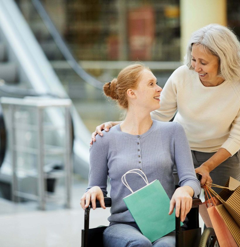 Young disabled woman in wheelchair talking to the nurse and smiling while they doing shopping together in the shopping mall